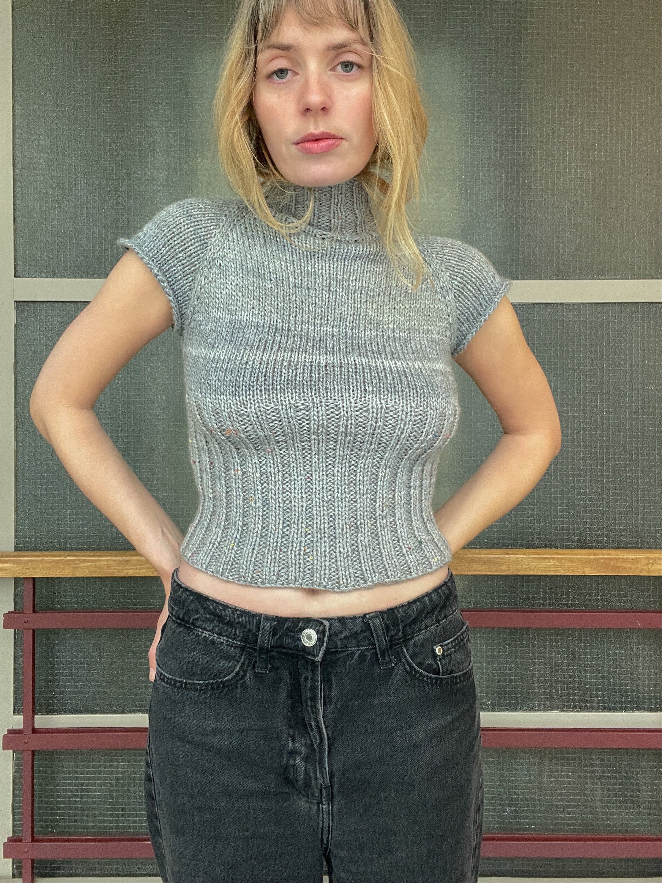Woman wearing a textured gray top and dark jeans standing against a screen door.